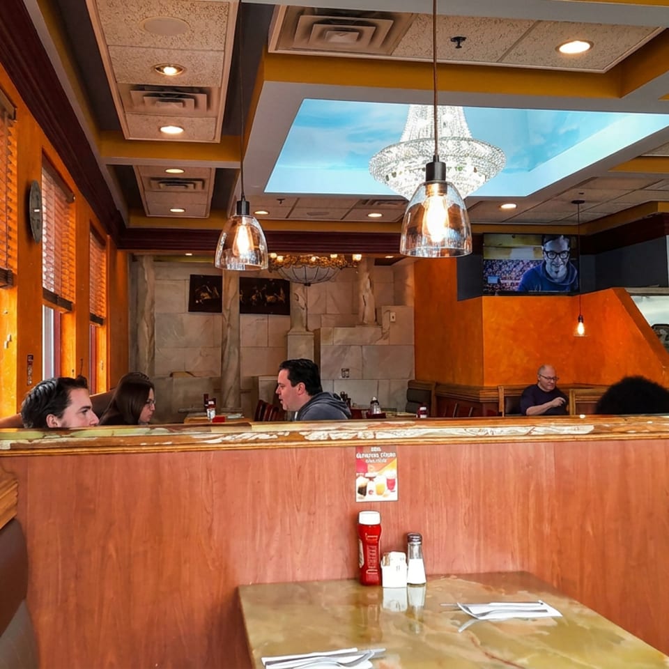Warmly lit restaurant dining room with orange walls, booths, pendant lights, seated diners, and an open kitchen.