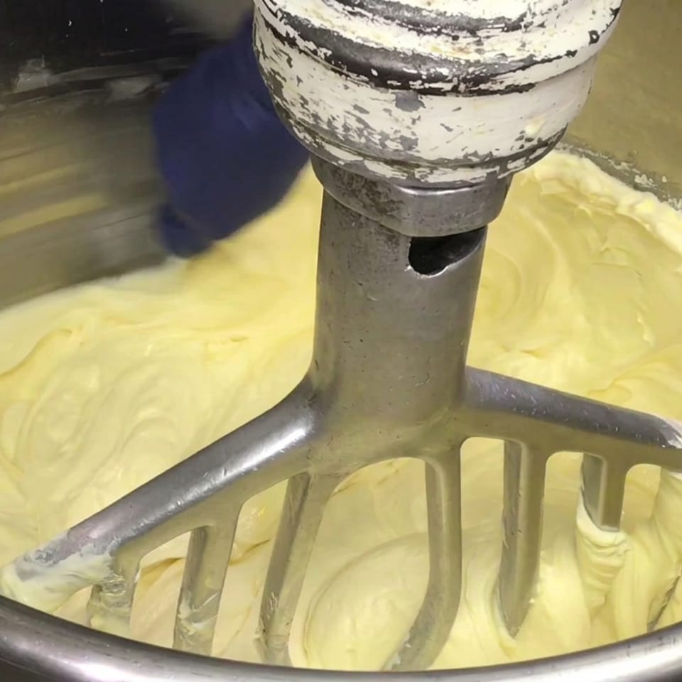 Industrial mixer paddle blending pale yellow batter in a large stainless steel bowl.